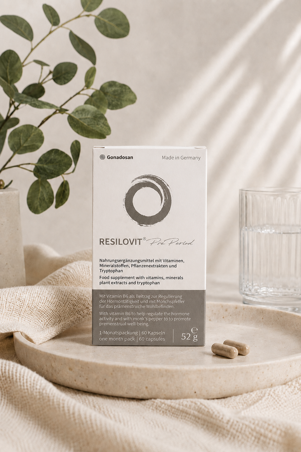Resilovit® Pre Period – Συμπλήρωμα διατροφής για υποστήριξη πριν από την περίοδο - Resilovit® Συσκευασία του Resilovit® Pre Period σε minimal σκηνικό με φυσικές αποχρώσεις, δίπλα σε ποτήρι νερού, φυλλώματα και κάψουλες, αναδεικνύοντας τη σύνθεσή του με βιταμίνες, μέταλλα, φυτικά εκχυλίσματα και τρυπτοφάνη.