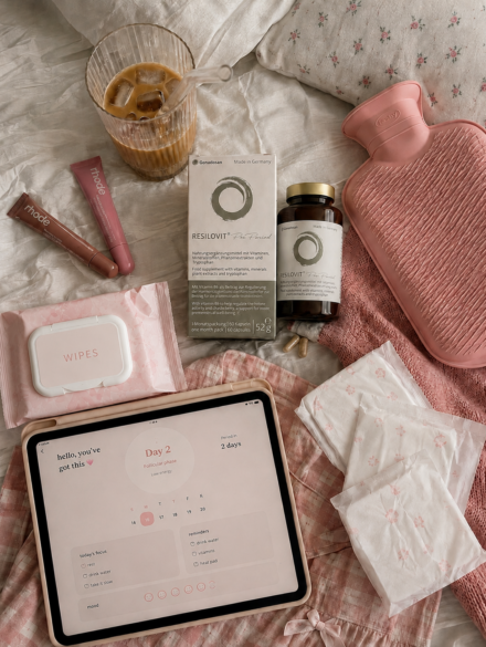 Flat lay of pink wellness items on a bed: water bottle with ice, supplements, wipes, a pink hot-water bottle, and a tablet showing a 'Day 2' plan.