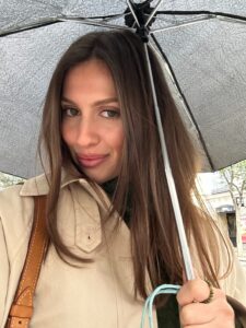 Woman with long brown hair under a large umbrella, wearing a beige trench coat and a brown leather shoulder bag visible at the left.