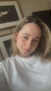 Close-up portrait of a woman with blond hair in a white shirt, wearing a delicate necklace, smiling softly indoors with framed photos in the background.