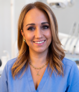 Smiling woman in blue scrubs wearing a gold chain necklace in a clinical exam room with dental equipment in the background